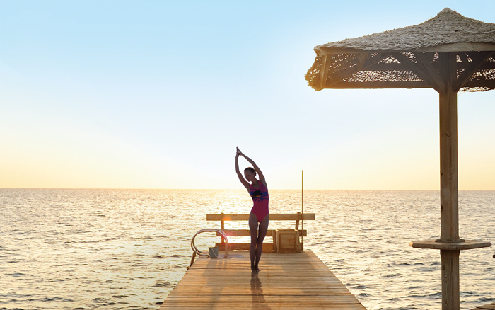 Woman practicing yoga on a wooden pier by the sea at sunrise – relaxation at The Breakers Somabay.